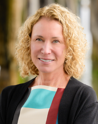 Smiling blonde woman in a black cardigan, holding a colorful folder against a blurred outdoor background