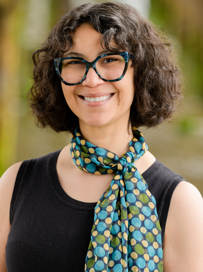 Smiling person with curly hair and glasses wearing a blue patterned scarf outdoors