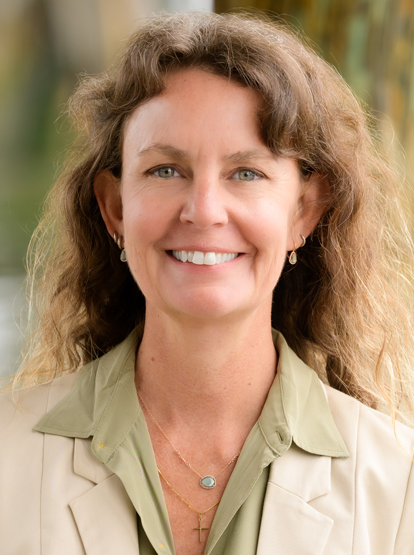 Smiling woman in a beige blazer and green blouse, standing outdoors with a blurred natural background