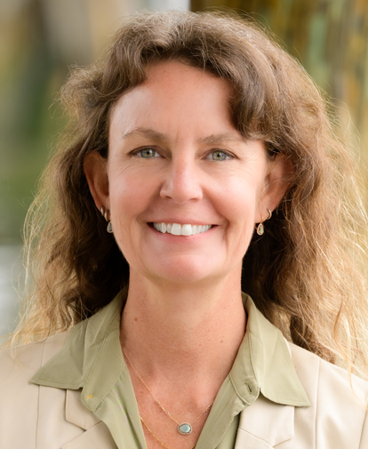 Smiling person with curly hair in a beige blazer and green shirt, standing outdoors with a blurred background
