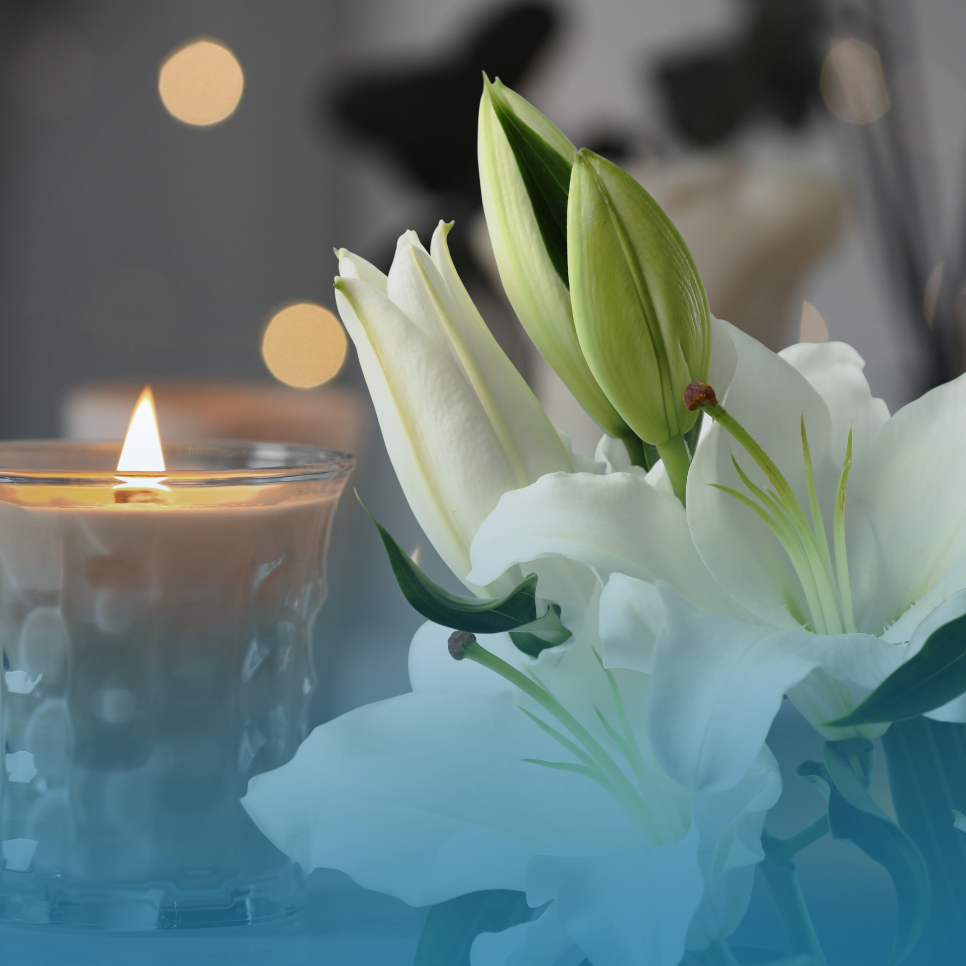 Lit candle beside white lilies with a soft blue haze and blurred bokeh lights in the background