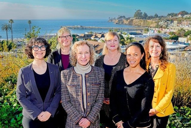 Six smiling people standing outdoors with ocean and coastal homes in the background