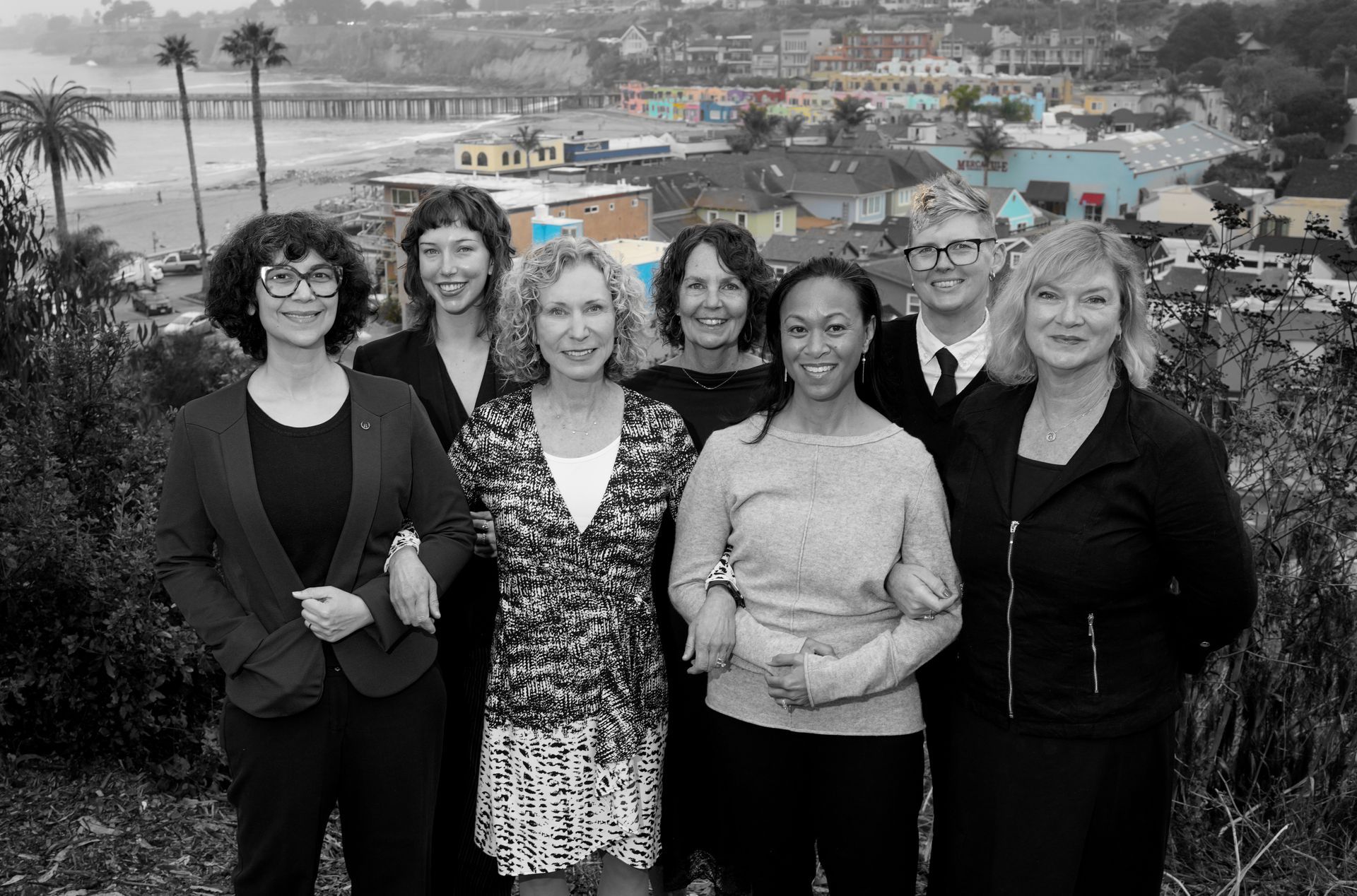 Six people standing outdoors by a coastline, with a city and bridge in the background.