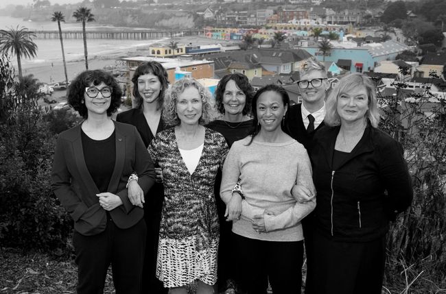 Six smiling people standing outdoors with ocean and coastal homes in the background