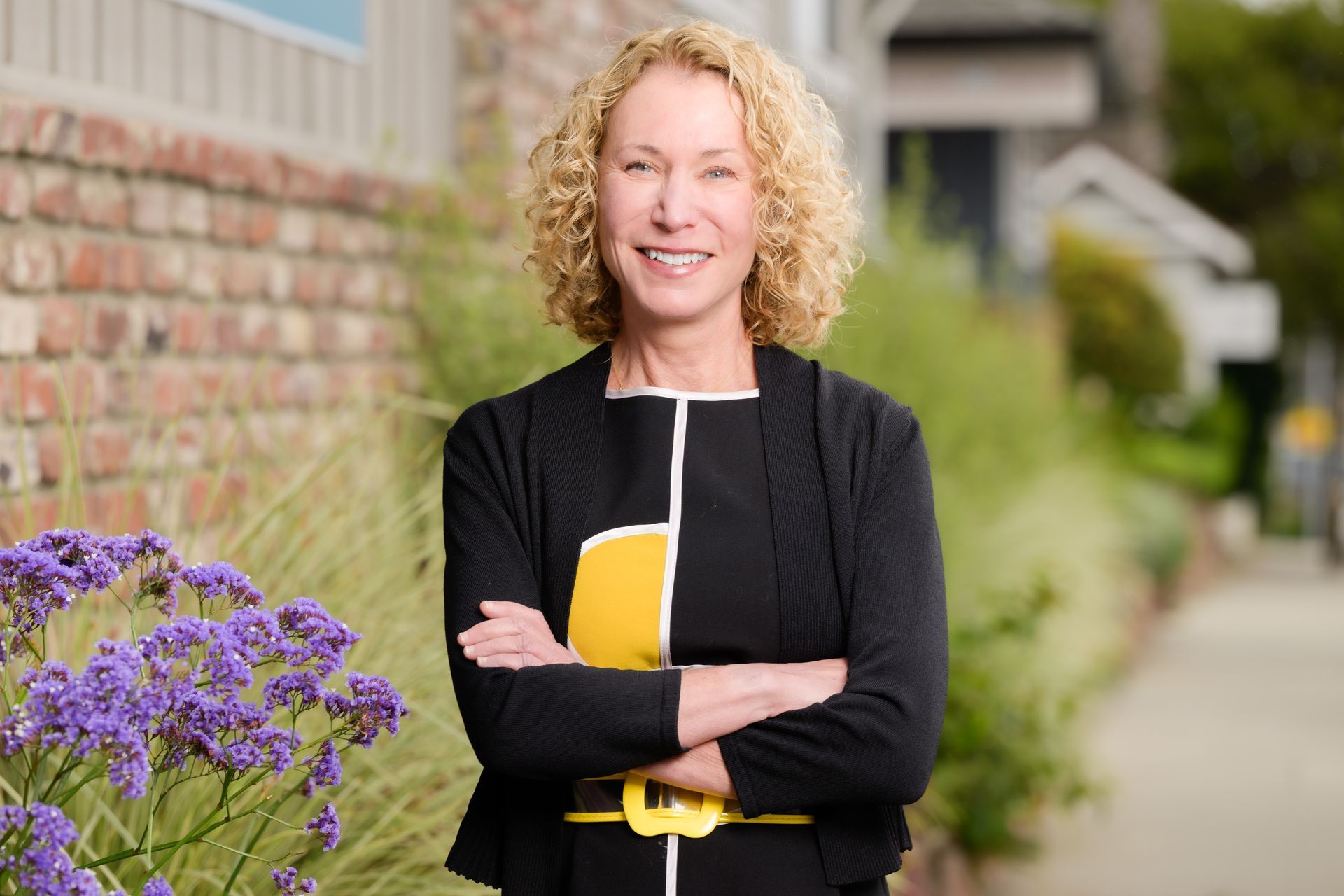 Woman standing with arms crossed beside purple flowers, outdoors near a brick building.