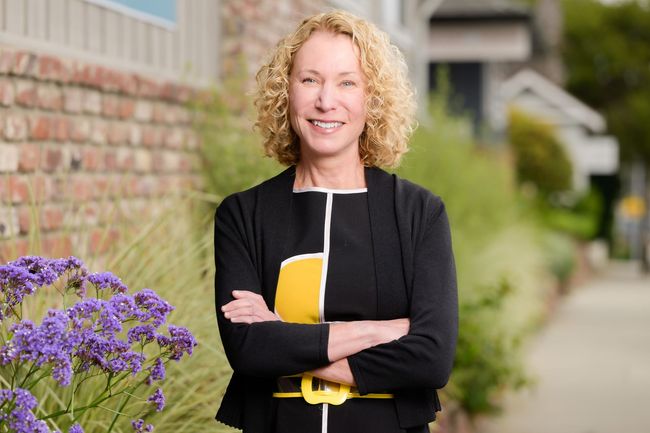 Woman standing with arms crossed beside purple flowers, outdoors near a brick building.