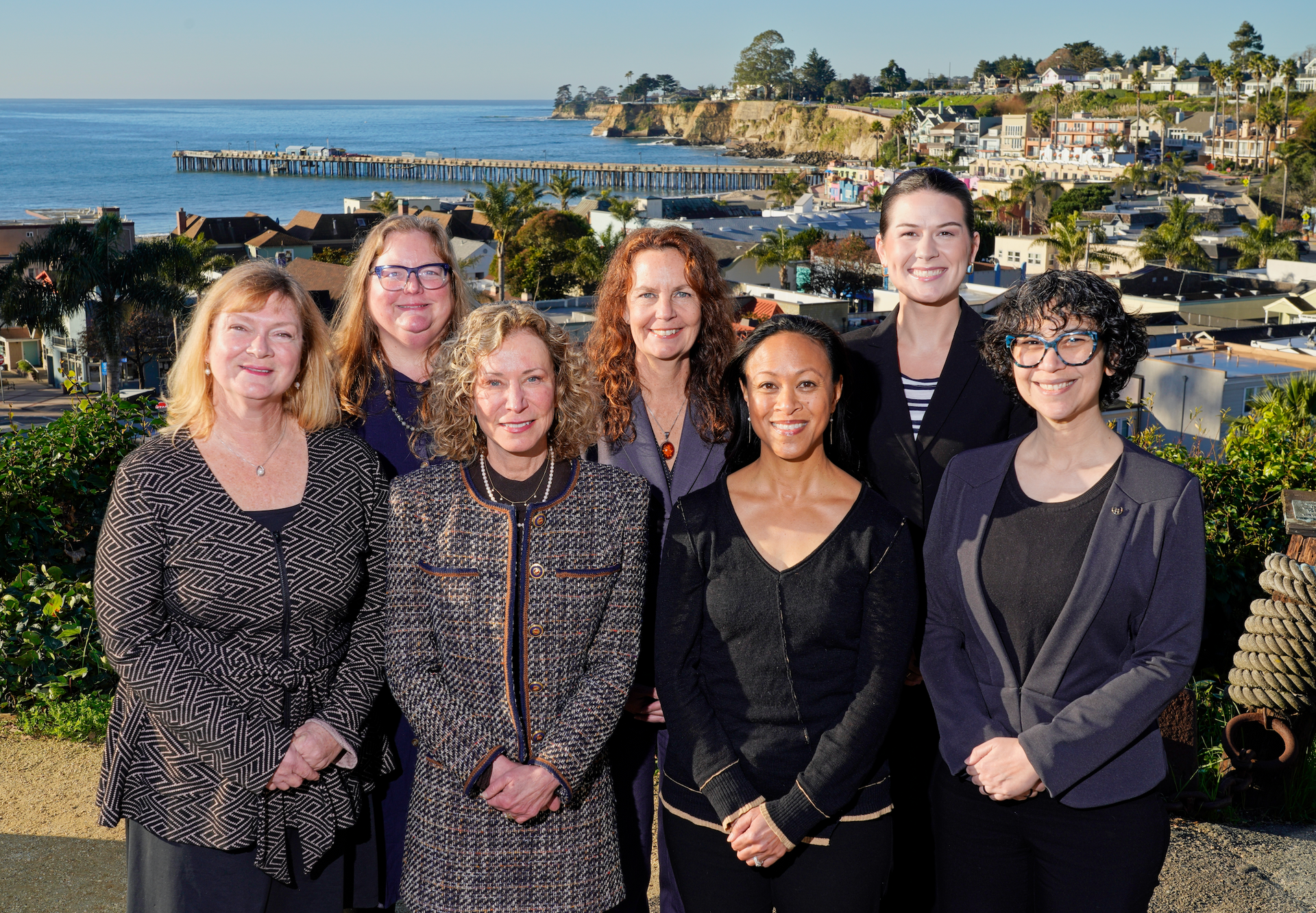 Six people posing outdoors with ocean and coastline in the background