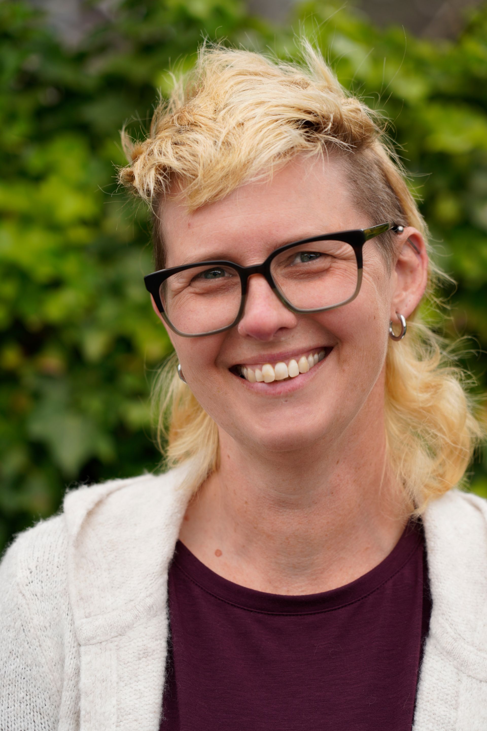 Smiling person with short blond hair and glasses outdoors, wearing a white cardigan and maroon shirt.