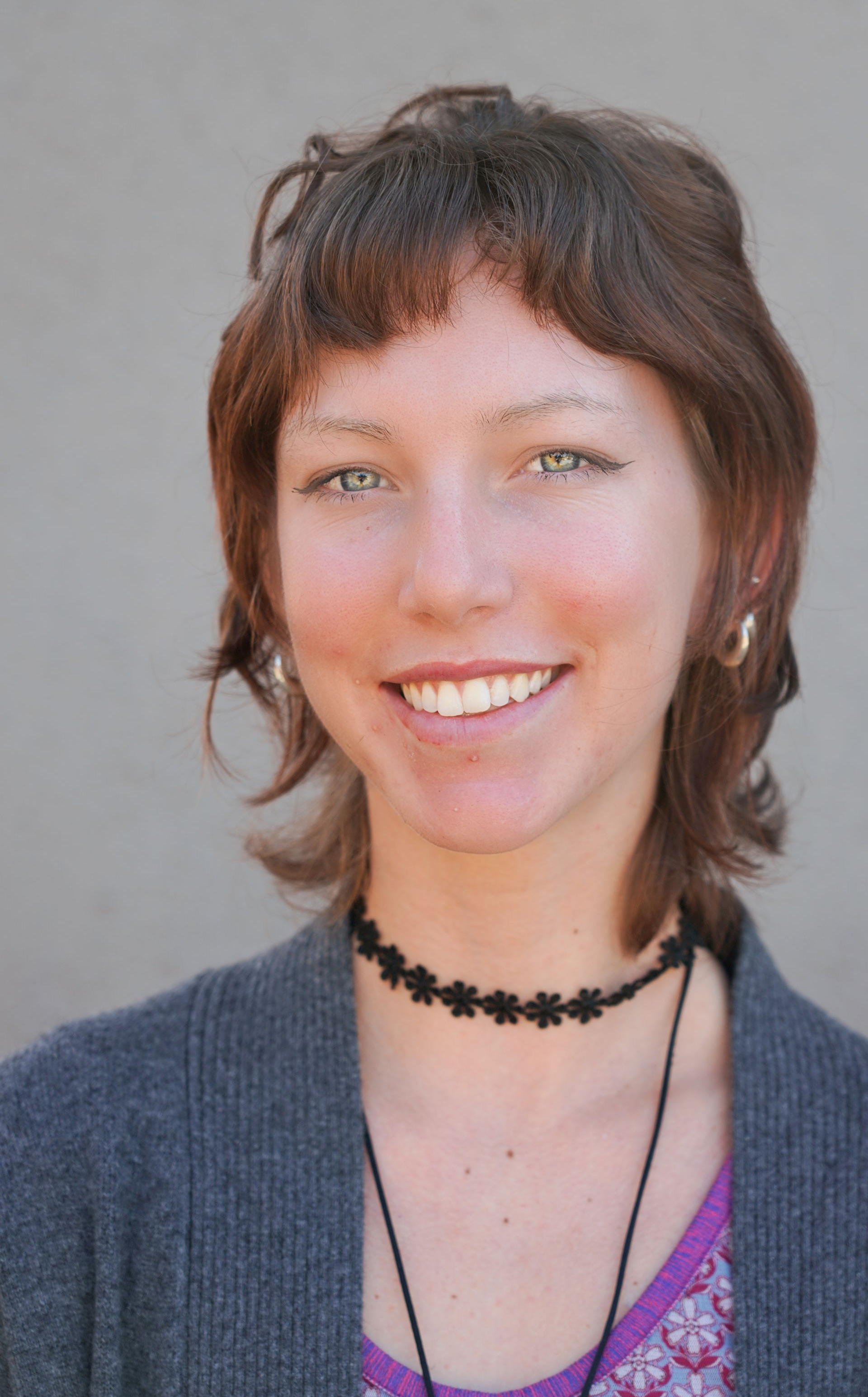 Portrait of a smiling person with short brown hair, wearing a gray cardigan and black choker necklace.