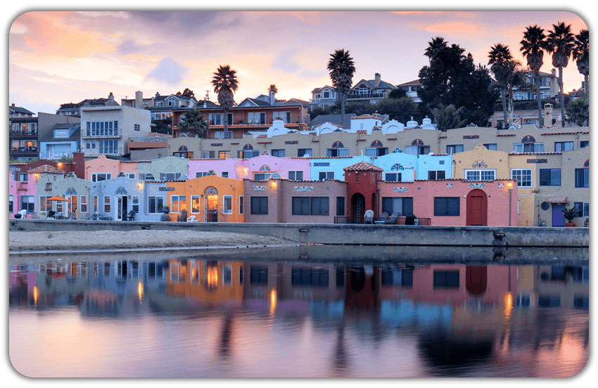 Pastel waterfront village in Capitola at sunset, with colorful buildings and palm trees reflecting in calm water.