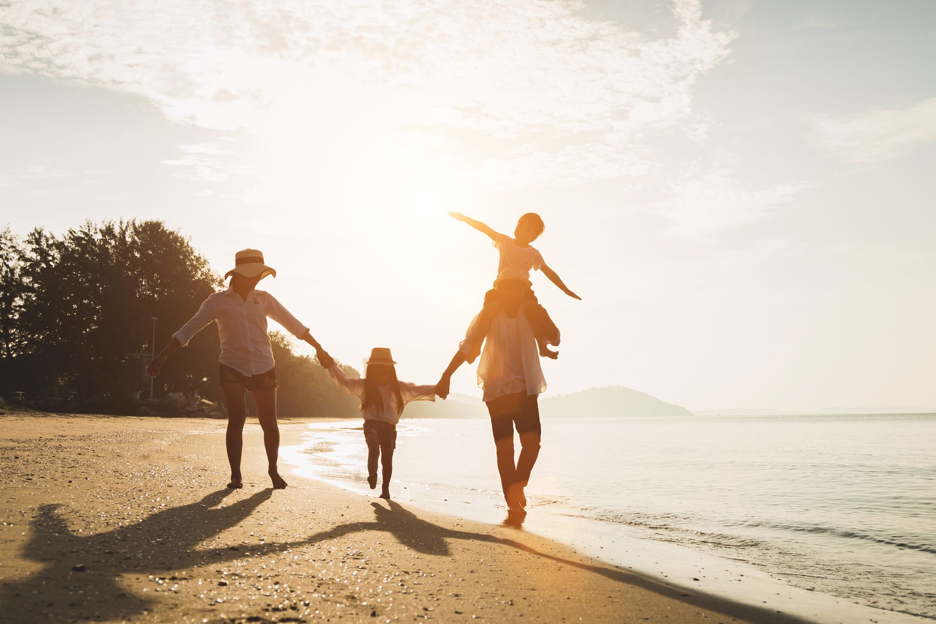 Family walking hand in hand along a beach at sunset, with a child between two adults and warm backlight.