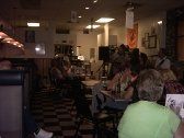 Restaurant interior with people seated at tables, listening to a performance. Dim lighting, checkered floor.