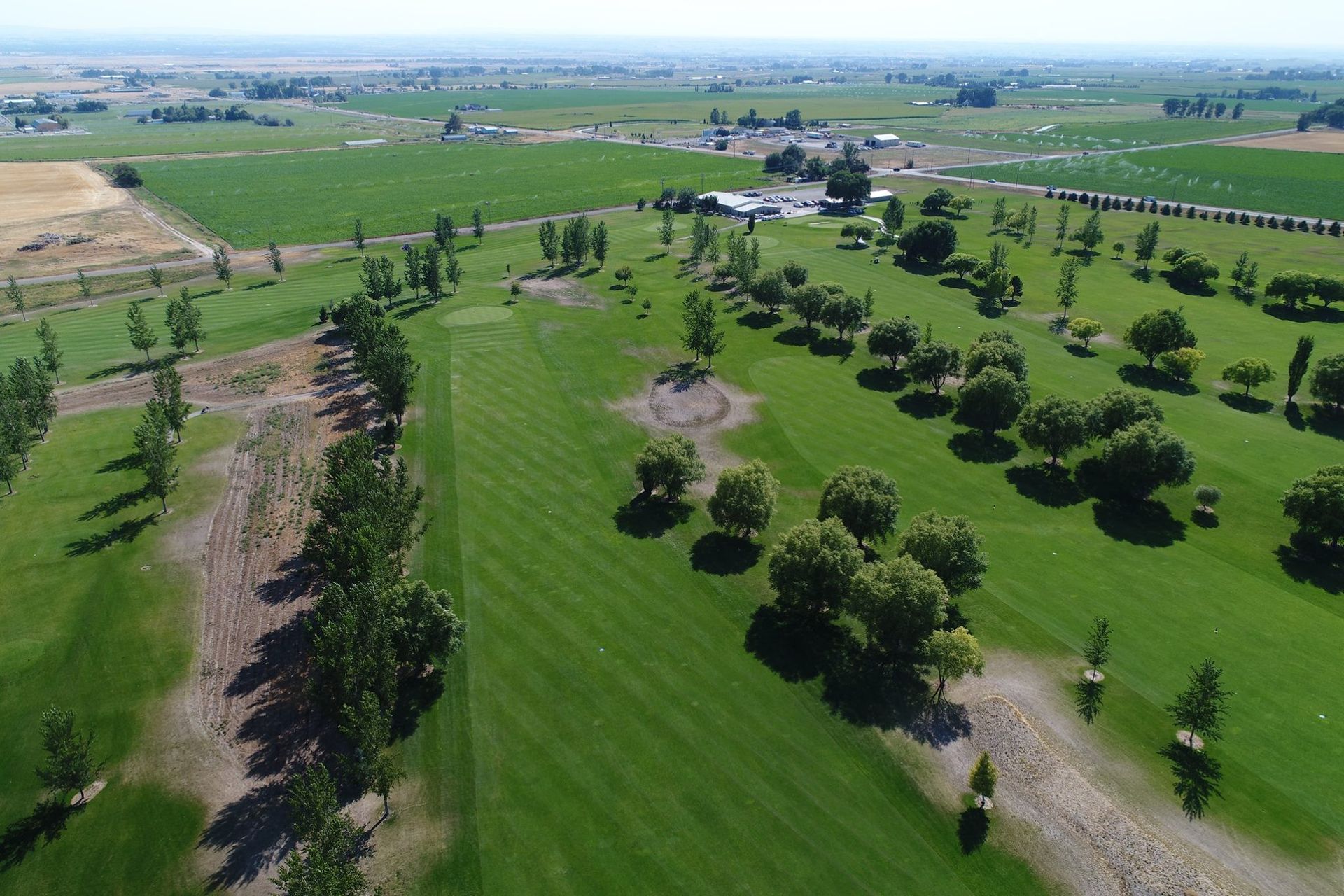 Aerial view of a green golf course with trees, fairways, and distant farmland under a clear sky