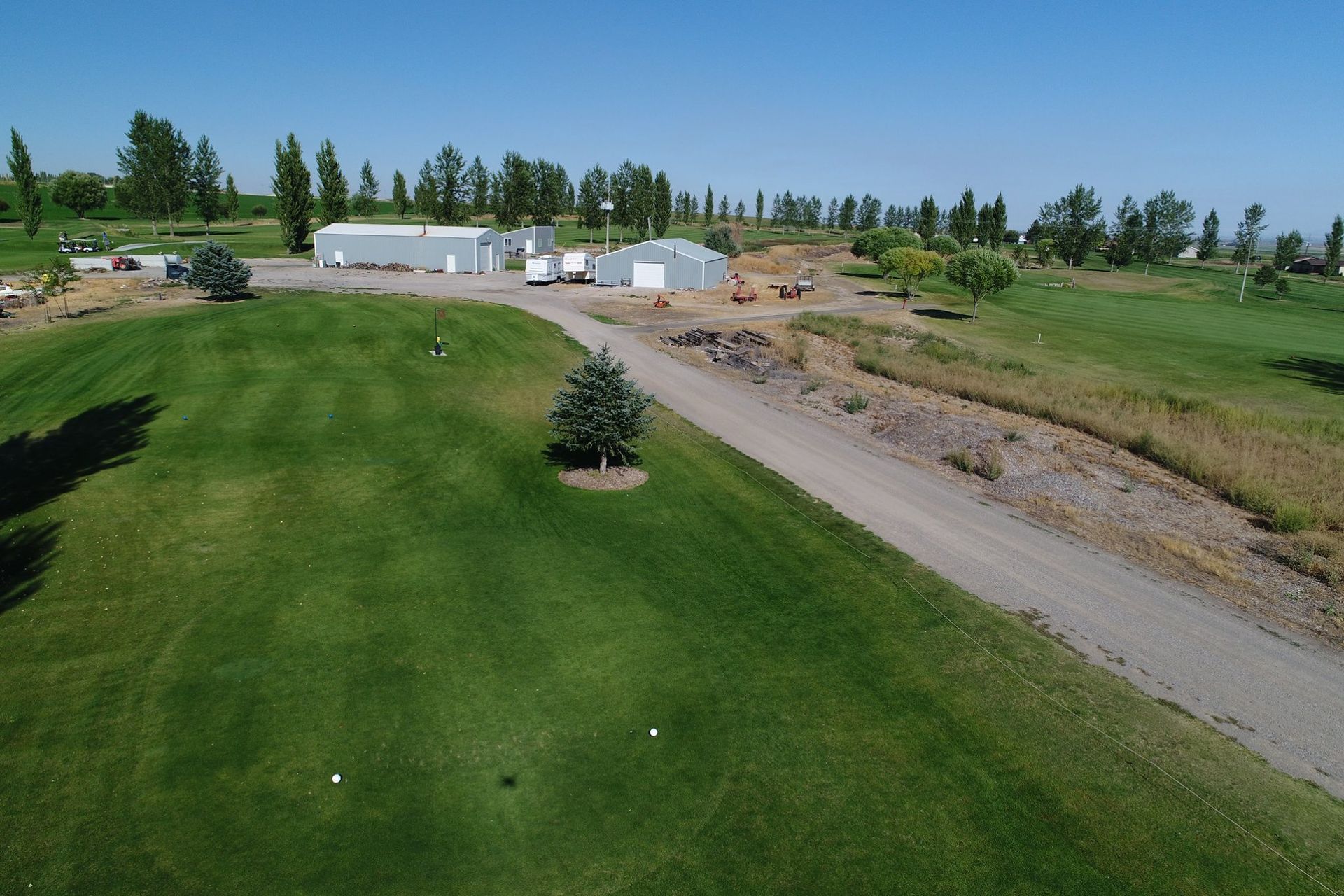 Aerial view of a green golf course beside a gravel road and white farm buildings under a blue sky