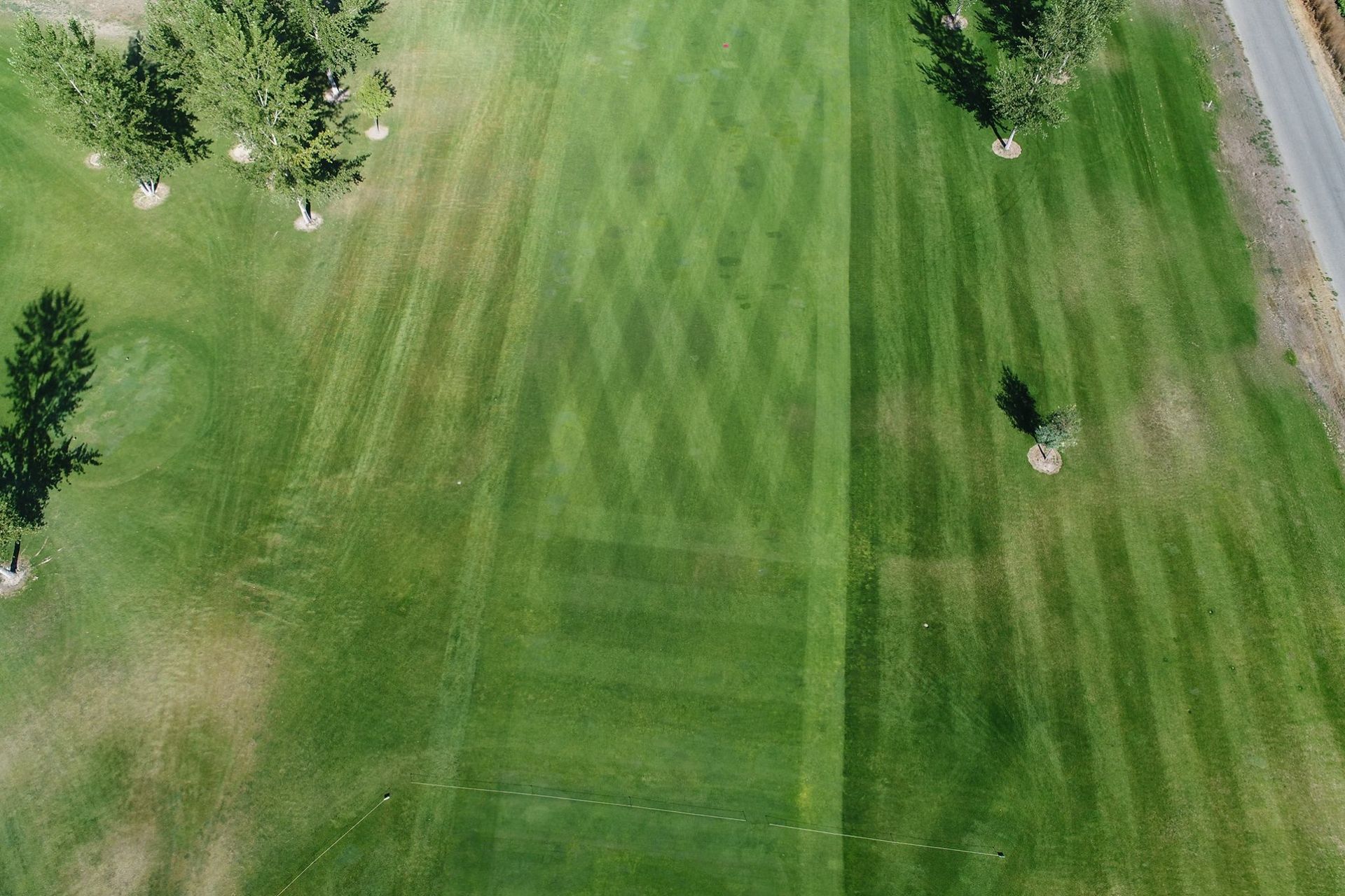 Aerial view of a green, striped golf fairway with trees and a path along the right edge