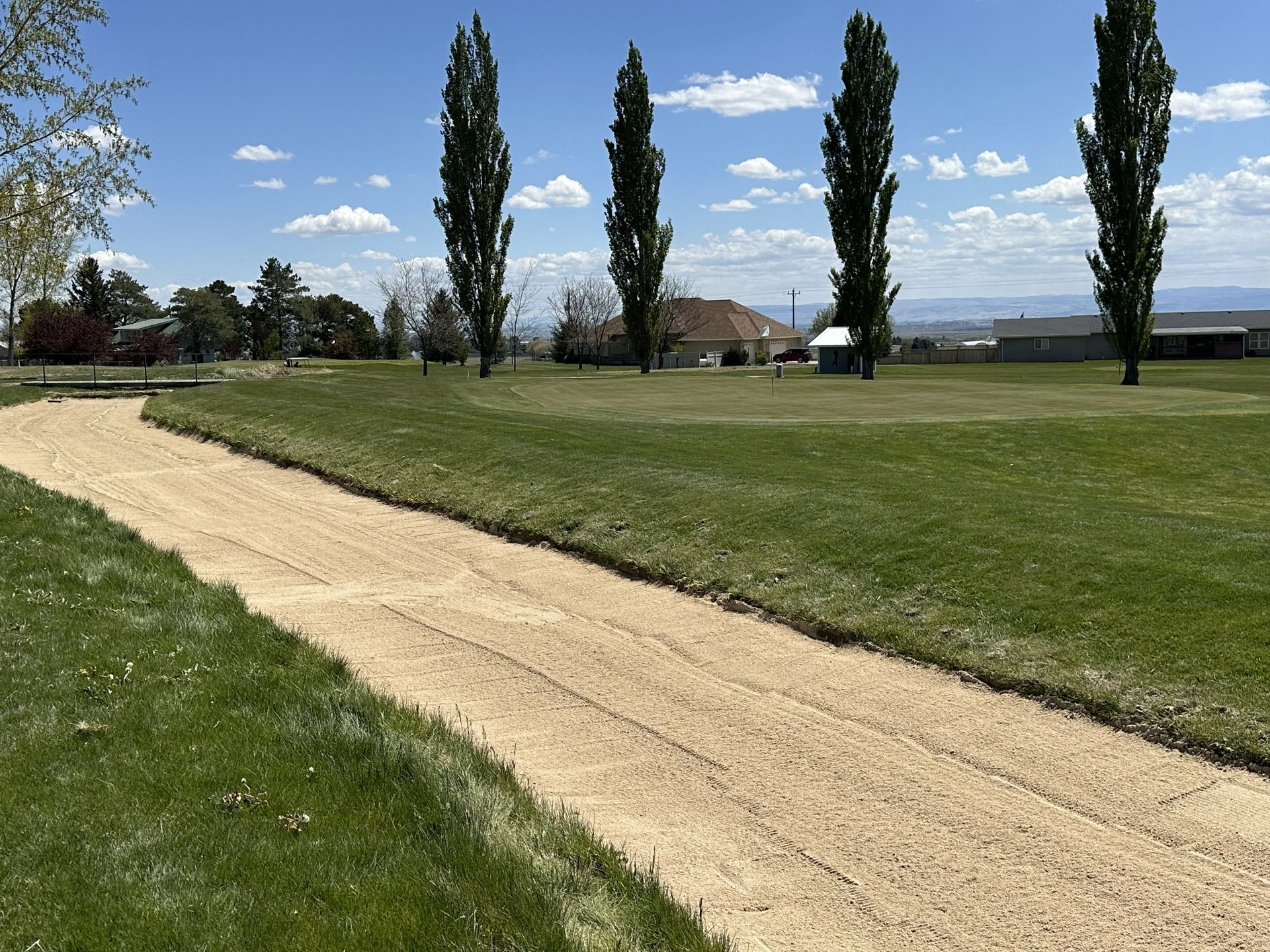 Gravel path through a green park with tall trees, under a blue sky with scattered clouds.