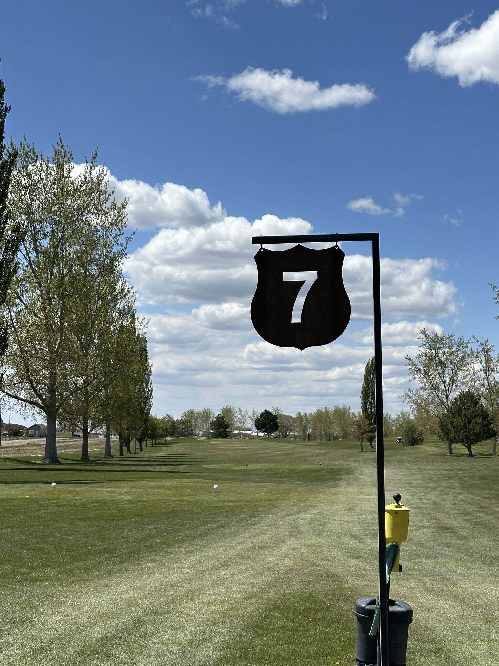 Golf course hole 7 sign on a cart path beside a grassy fairway under a blue sky with clouds