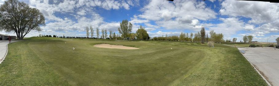 Wide grassy golf course with sand bunker, scattered trees, and a paved path under a partly cloudy sky