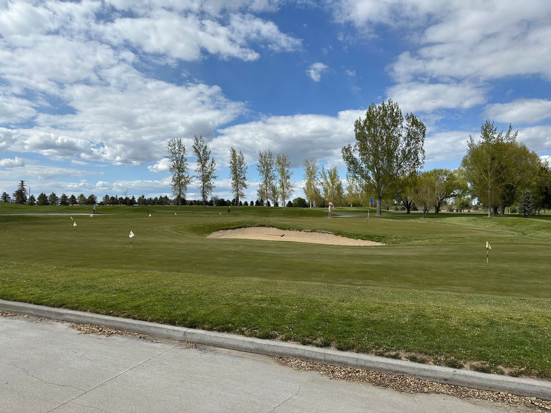 Golf course fairway with sand bunker, green grass, trees, and a cloudy blue sky.