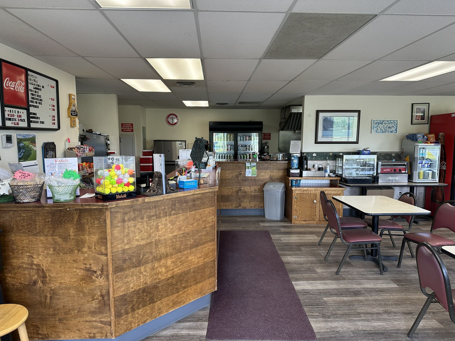 Small café lobby with a curved wooden counter, tables, chairs, and menu boards under bright ceiling lights.