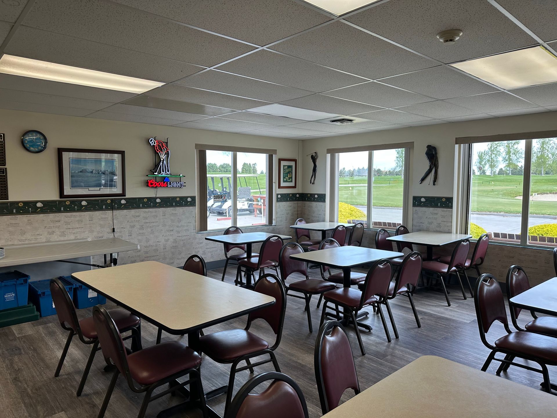 Empty community room with tables and chairs, windows overlooking grass, and a kitchen area along the wall