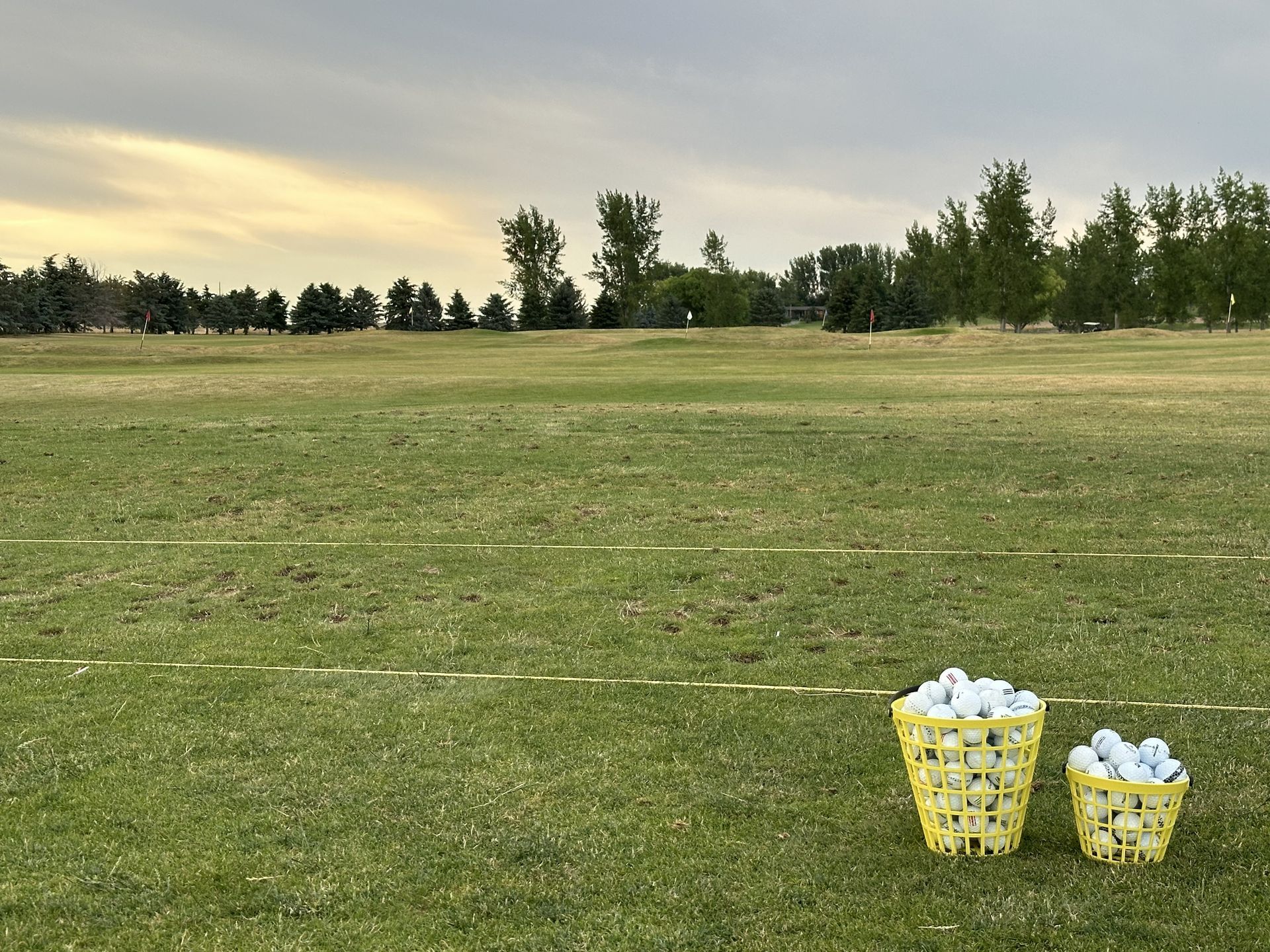 Two yellow baskets of white balls on a grassy field under a cloudy sky