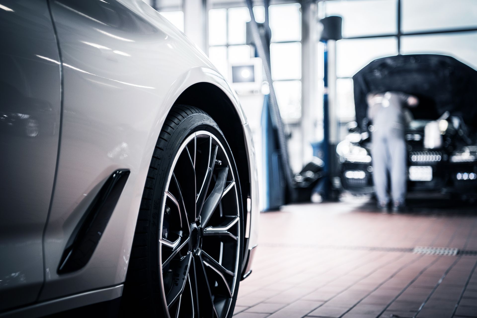 Silver car tire in a repair shop with a mechanic working on another car in the background. | Rocky Mountain Car Care
