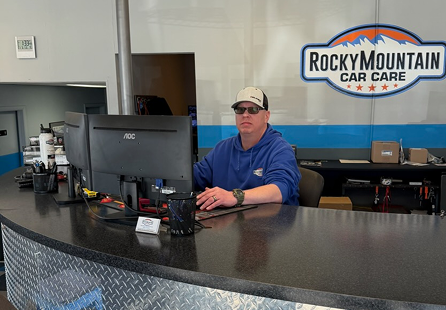 Man at a computer in an auto repair shop. Rocky Mountain Car Care sign in the background. | Rocky Mountain Car Care