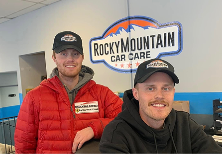 Two men in caps stand near a Rocky Mountain Car Care sign. One wears a red jacket, the other a black hoodie. | Rocky Mountain Car Care