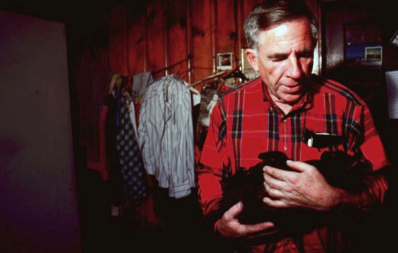 Man in red plaid shirt cradles a cat in his arms. He's standing in a dimly lit room with clothes hanging in the background.