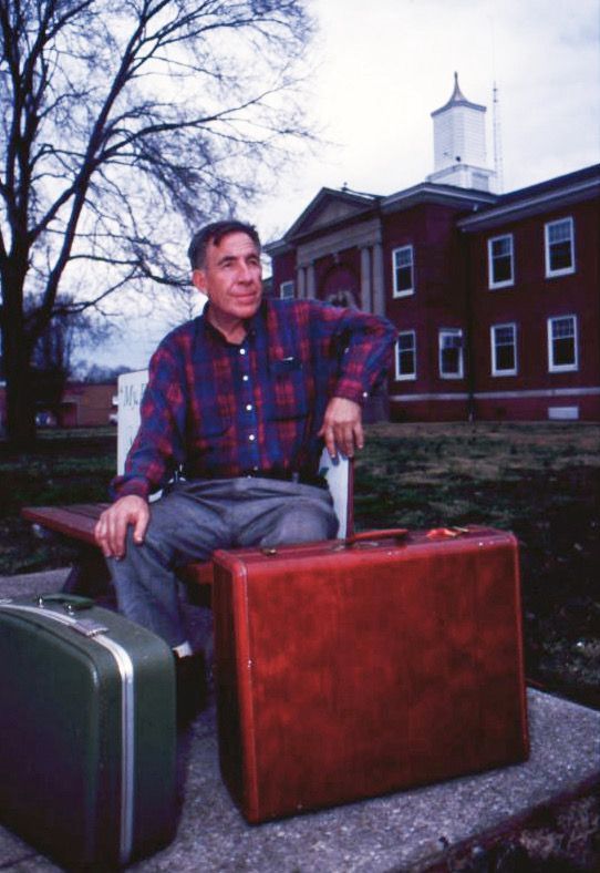 Man sits on a bench with two suitcases, outside a brick building. He's in a plaid shirt and jeans, looking pensively toward the sky.