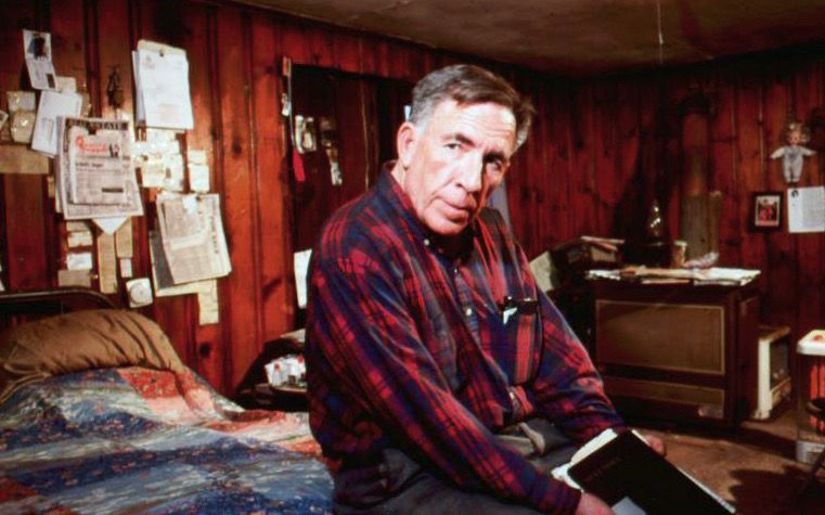 Man in a plaid shirt sits on a bed in a cluttered room. Red wood paneling, papers on the wall, and a book in hand.