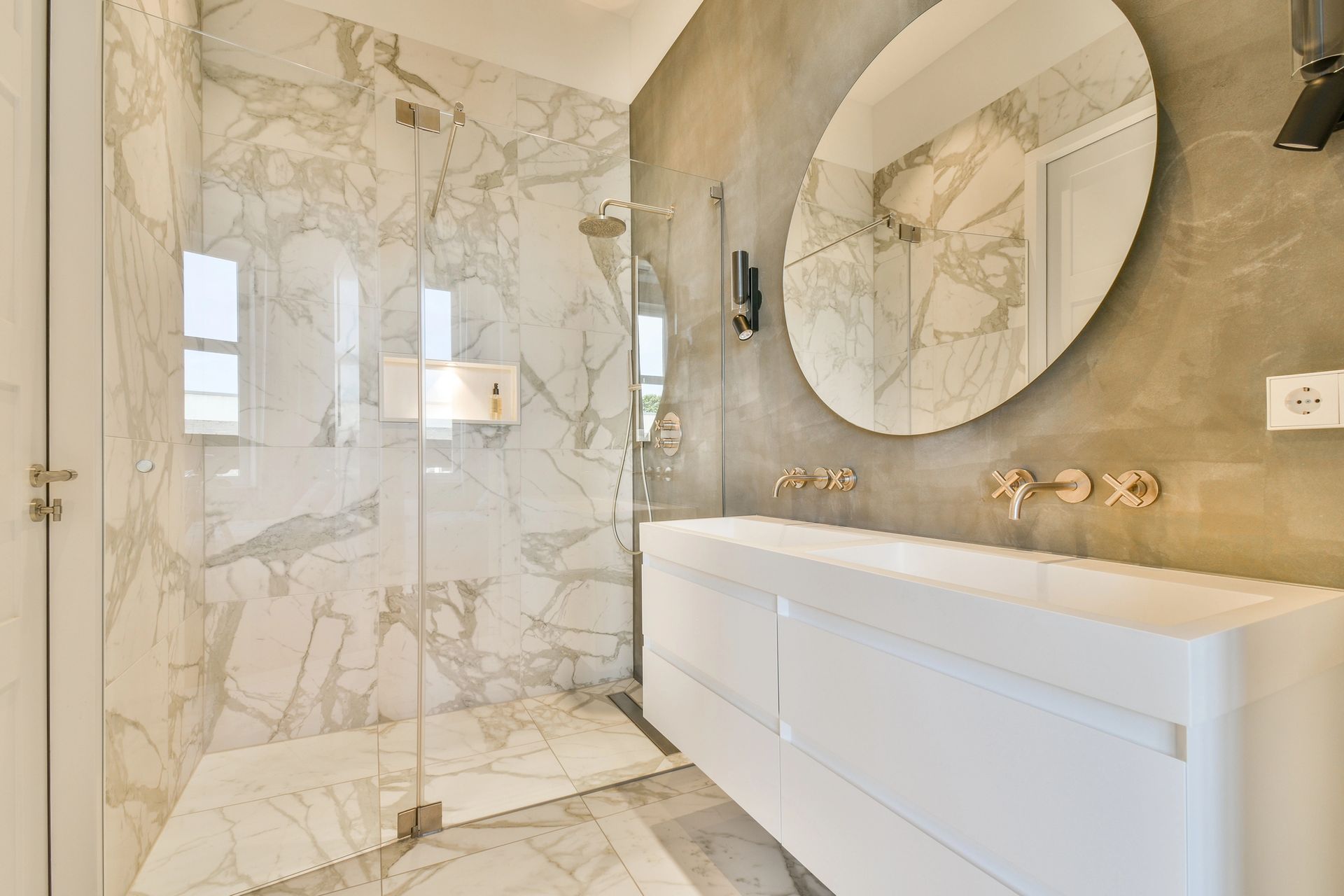Elegante baño de mármol con ducha a ras de suelo, espejo redondo y tocador blanco.