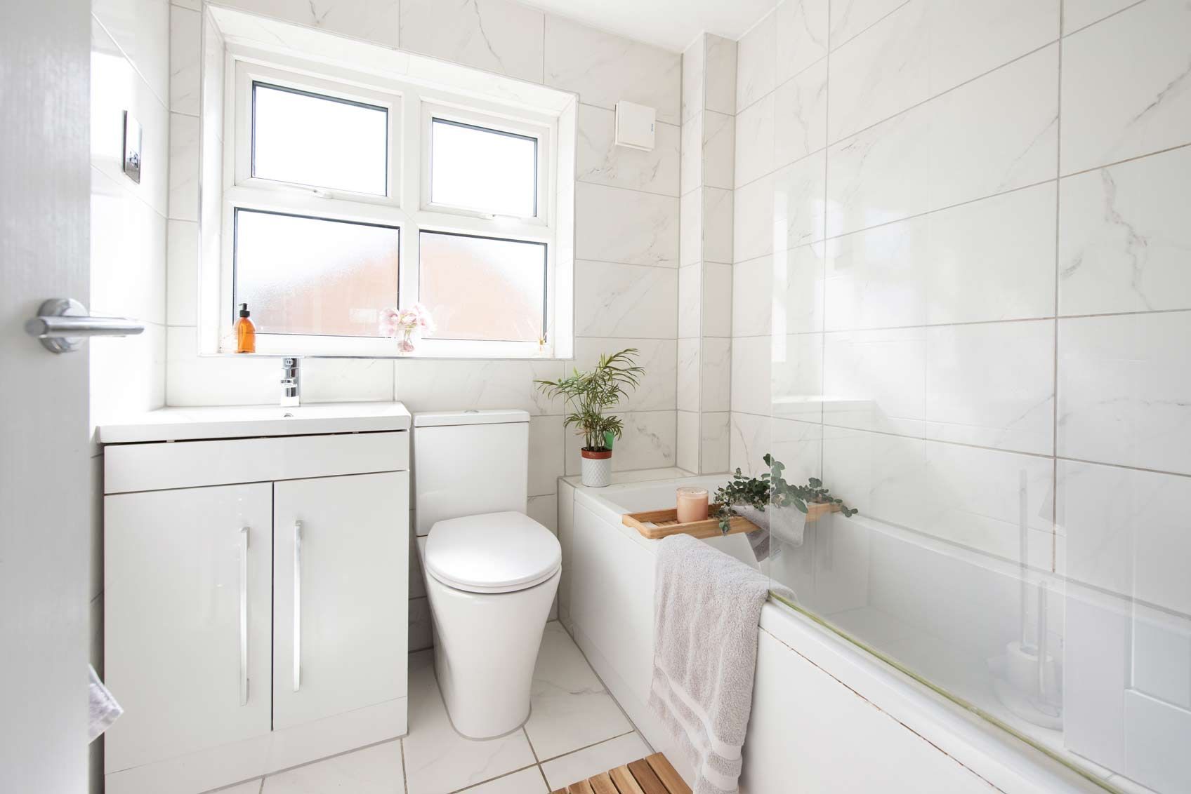 White tiled bathroom with sink, toilet, bathtub, and window.