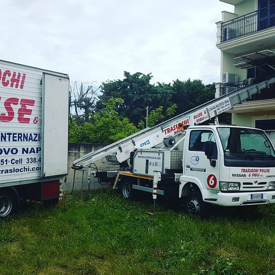 Un furgone bianco con scala pieghevole è parcheggiato sull'erba accanto a un edificio e a un camion più grande.