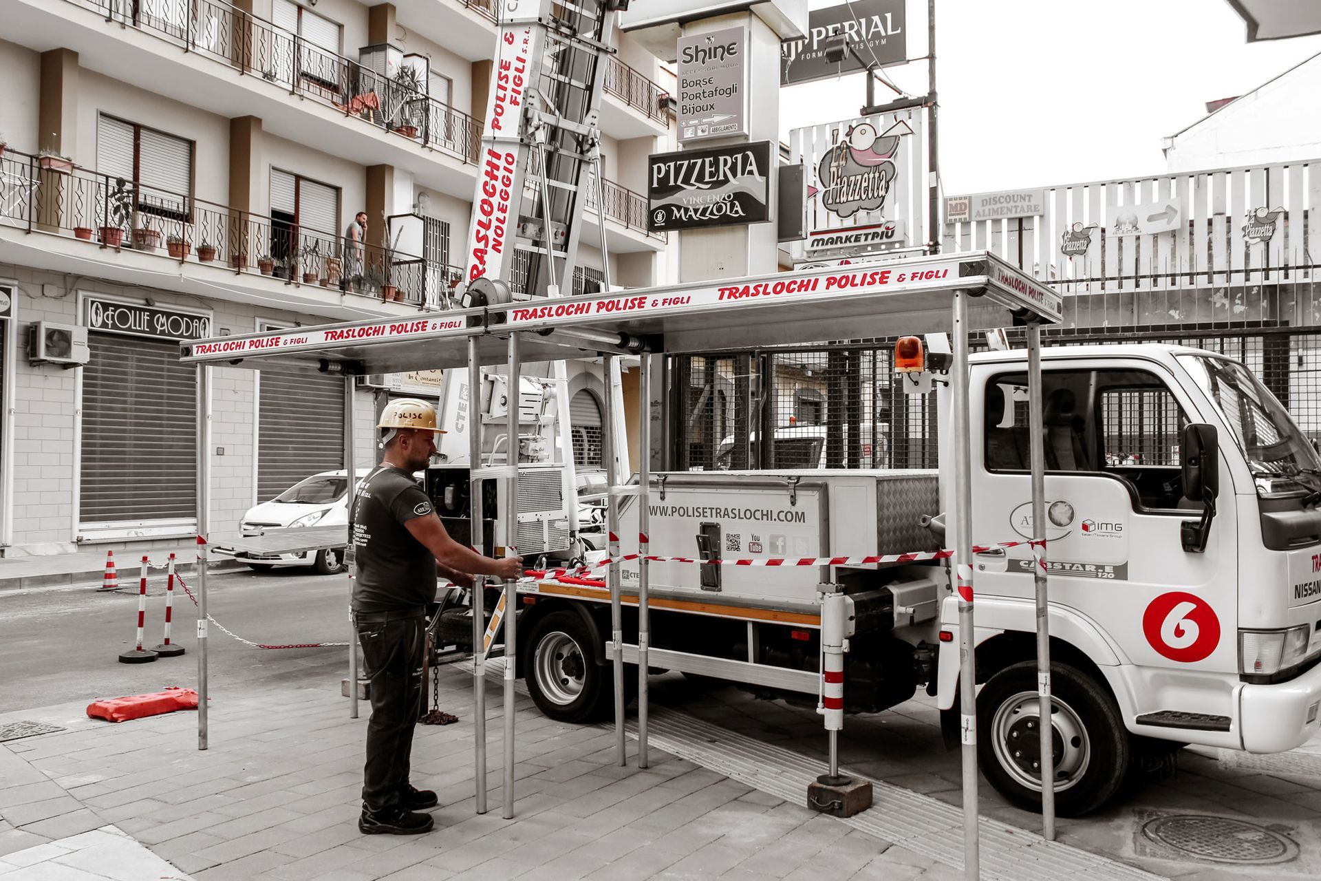Un operaio con l'elmetto guida un camioncino bianco di servizio all'interno di una strada chiusa da un cancello in una zona urbana.
