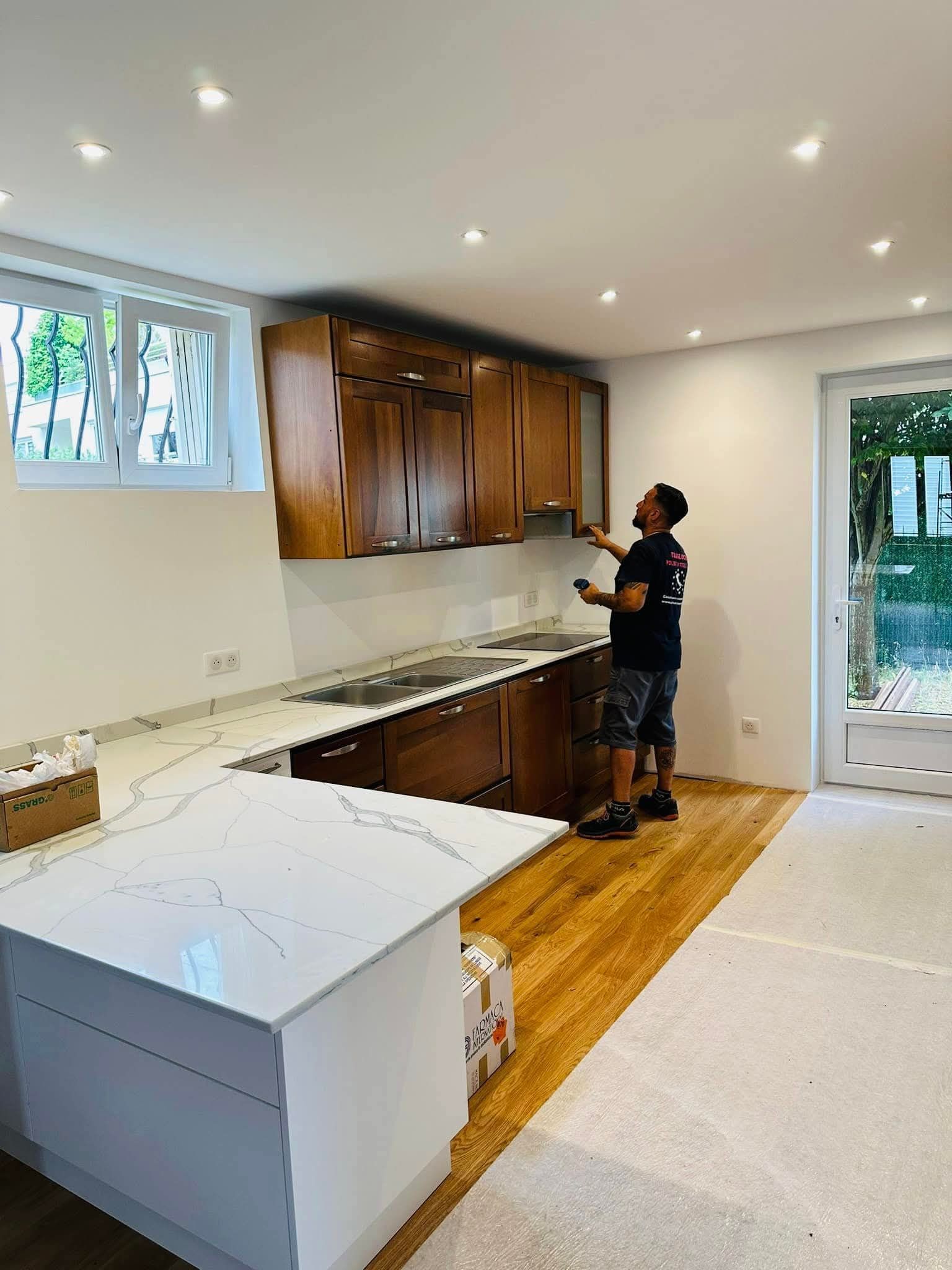 Un operaio installa dei mobili in una cucina luminosa e vuota con ripiani bianchi e pavimento in legno.