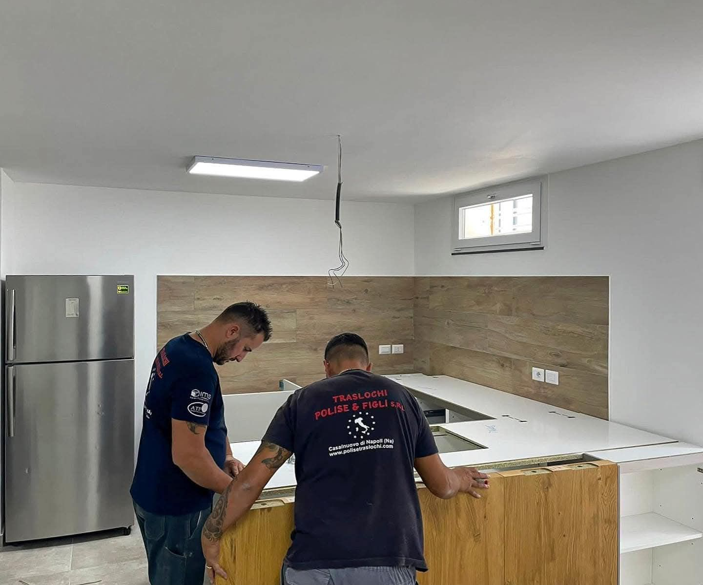 Due operai installano un'isola cucina in una stanza luminosa e non rifinita, dotata di frigorifero in acciaio inossidabile.