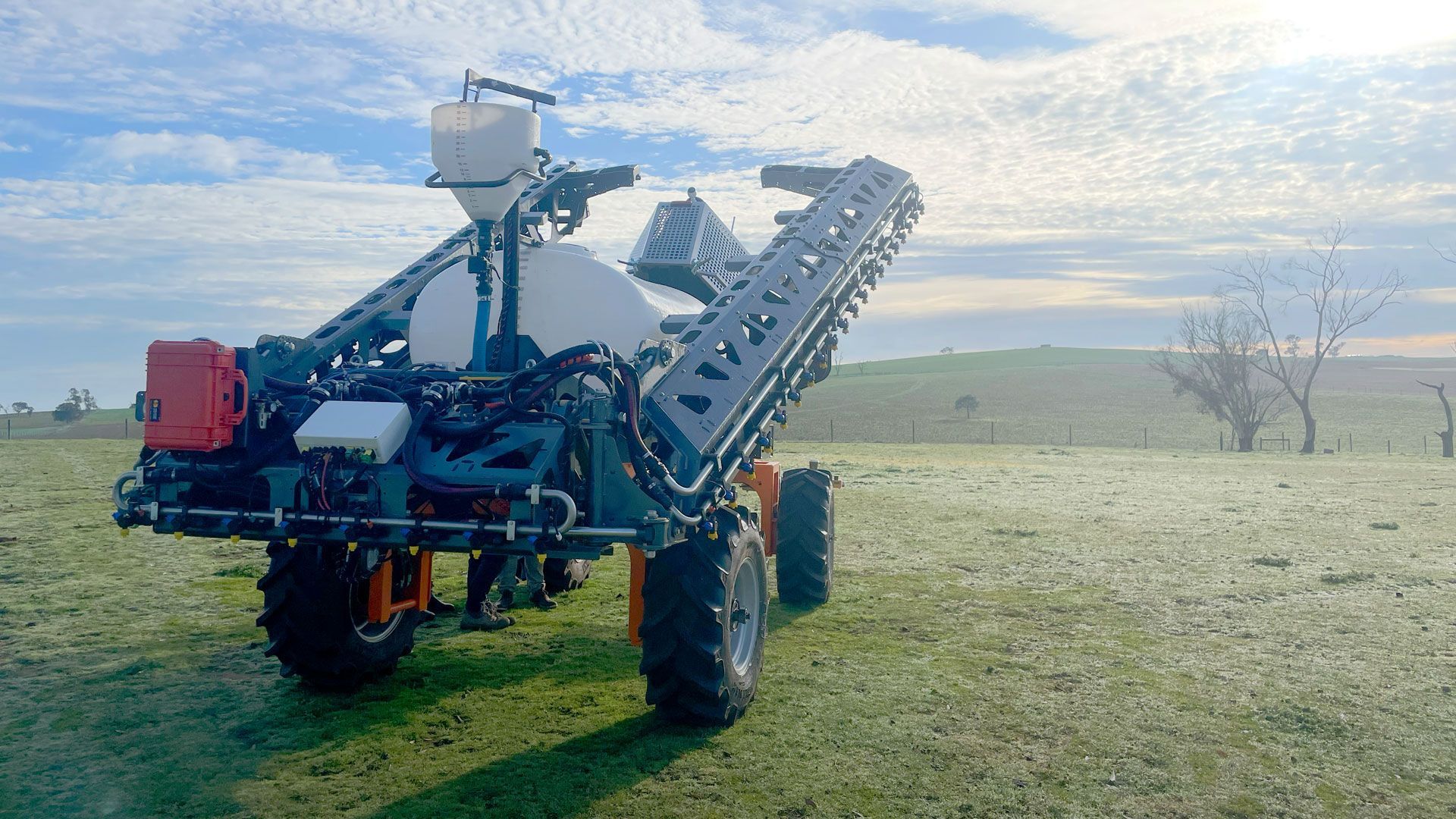 Automated hydraulic Agricultural sprayer in a field. The machine is orange and black, with a white tank. Sunny, outdoor setting.