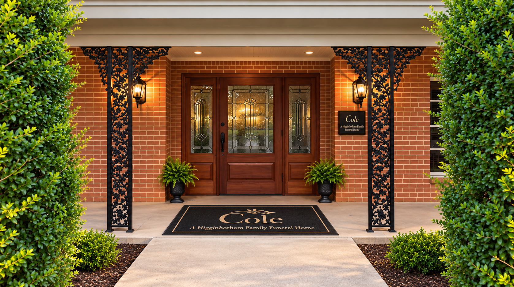 Brick house front entrance with double wooden doors, black lanterns, and a welcoming doormat surrounded by greenery