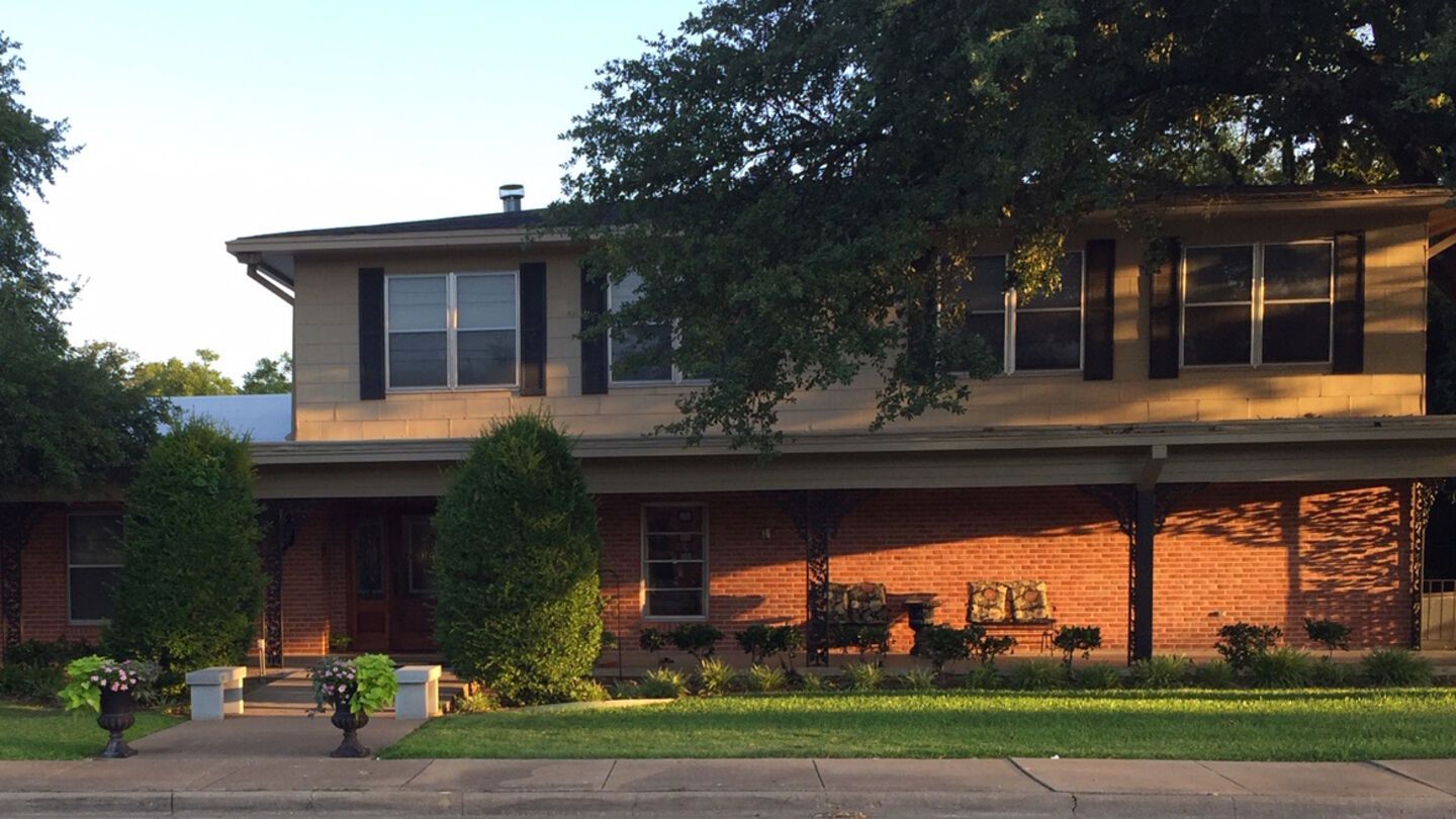 Two-story brick house with a front porch, trimmed shrubs, and a tree in the yard at dusk