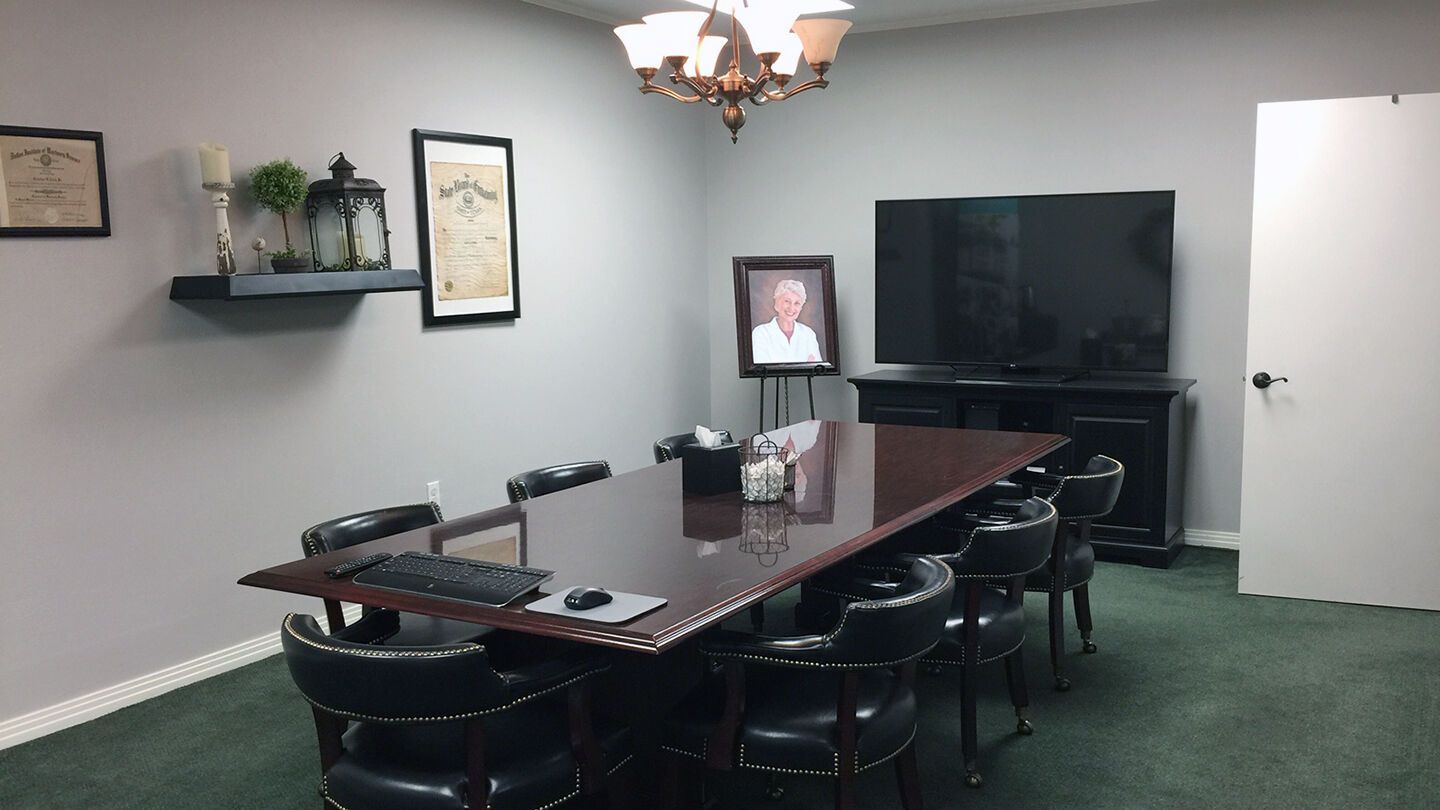Conference room with long table, black chairs, wall-mounted TV, framed portrait, and green carpet.