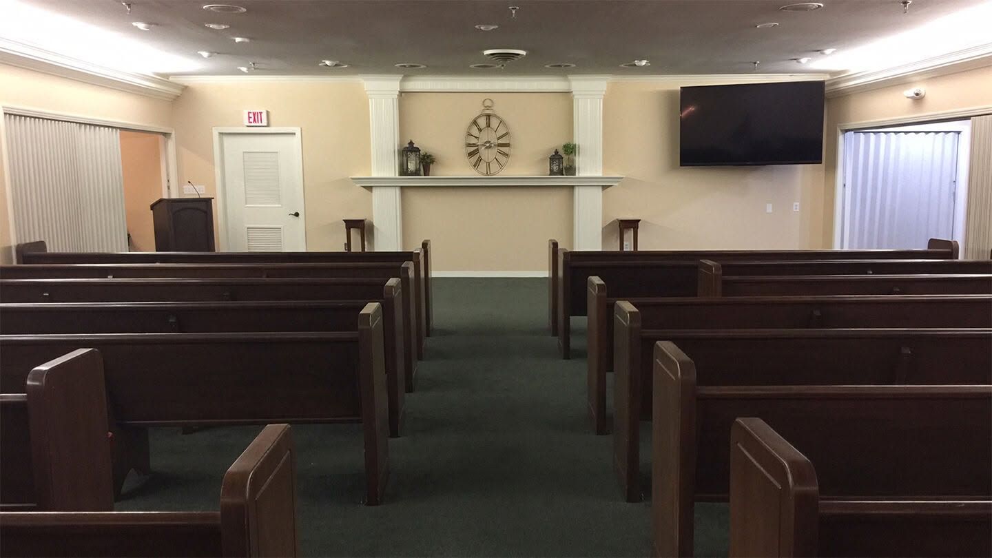 Small church sanctuary with wooden pews, green carpet, and a white altar wall with cross and TV screens