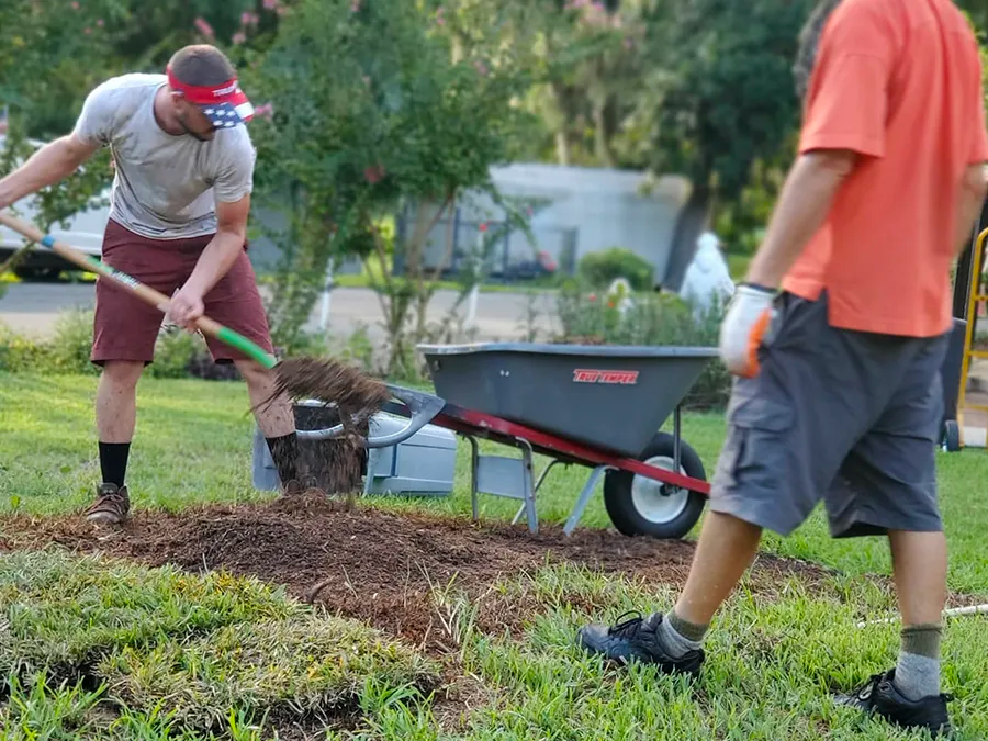 Two men are working in a yard with a wheelbarrow.