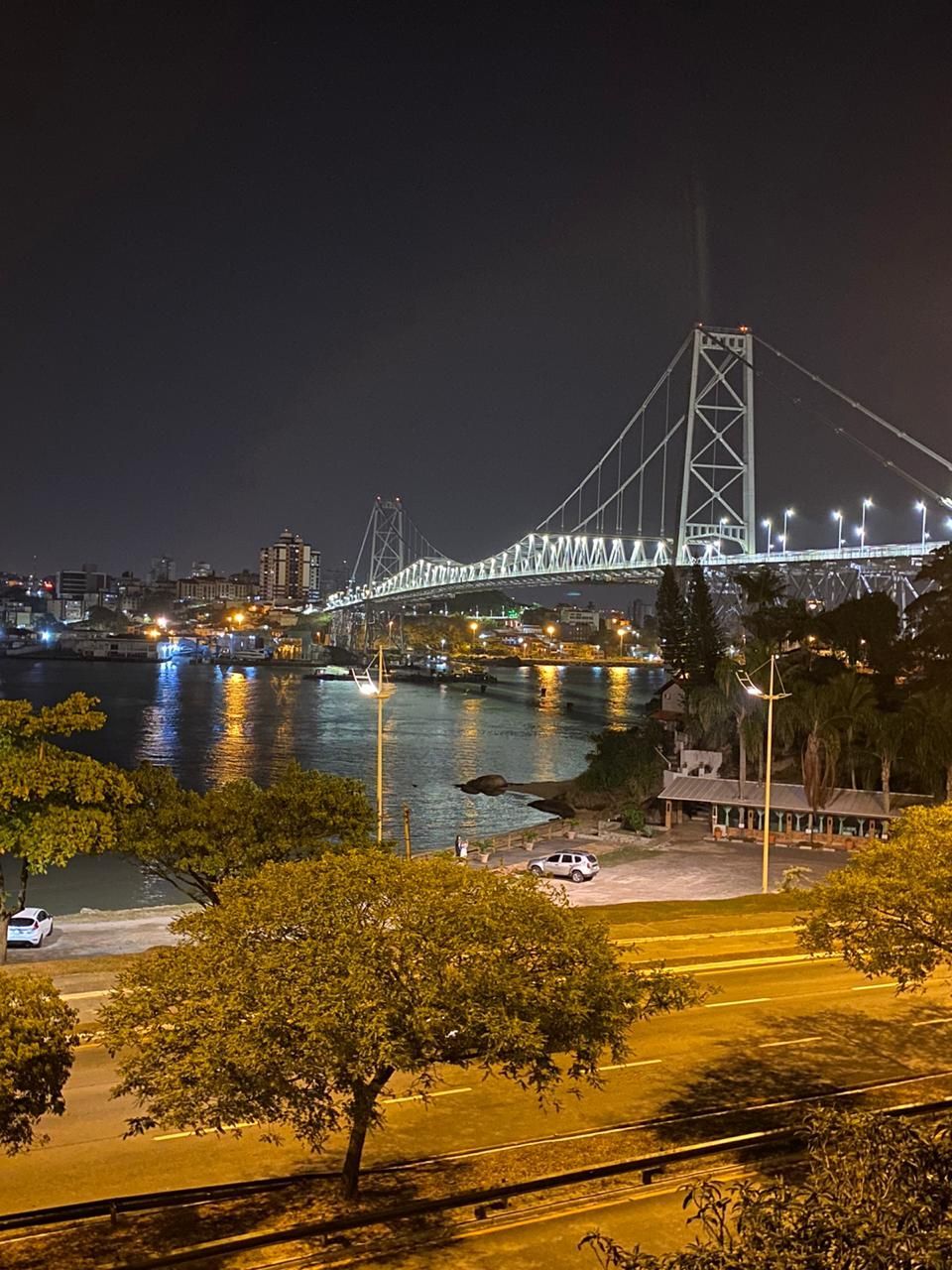 Vista noturna de uma ponte suspensa bem iluminada sobre um corpo d'água, com as luzes da cidade ao fundo.