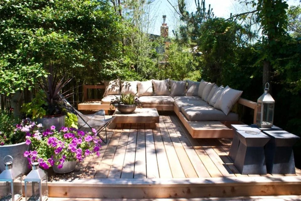 Patio esterno in legno con divano componibile, circondato da una vegetazione lussureggiante e fiori.
