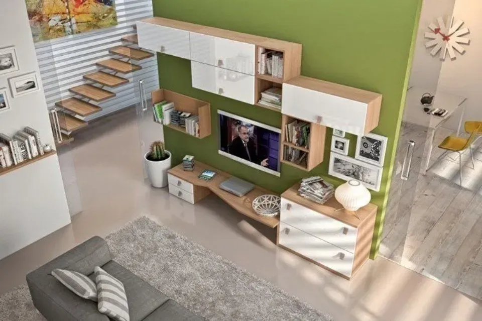 Soggiorno moderno con parete verde, TV e mobili in legno chiaro. Visibili anche un divano grigio, un tappeto e una scala.