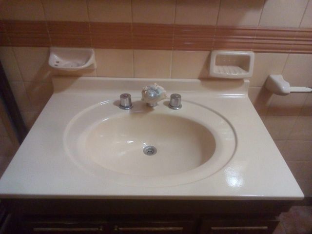 Lavabo de baño con encimera blanca, lavabo color crema, grifo plateado y dispensador de jabón montado en la pared.