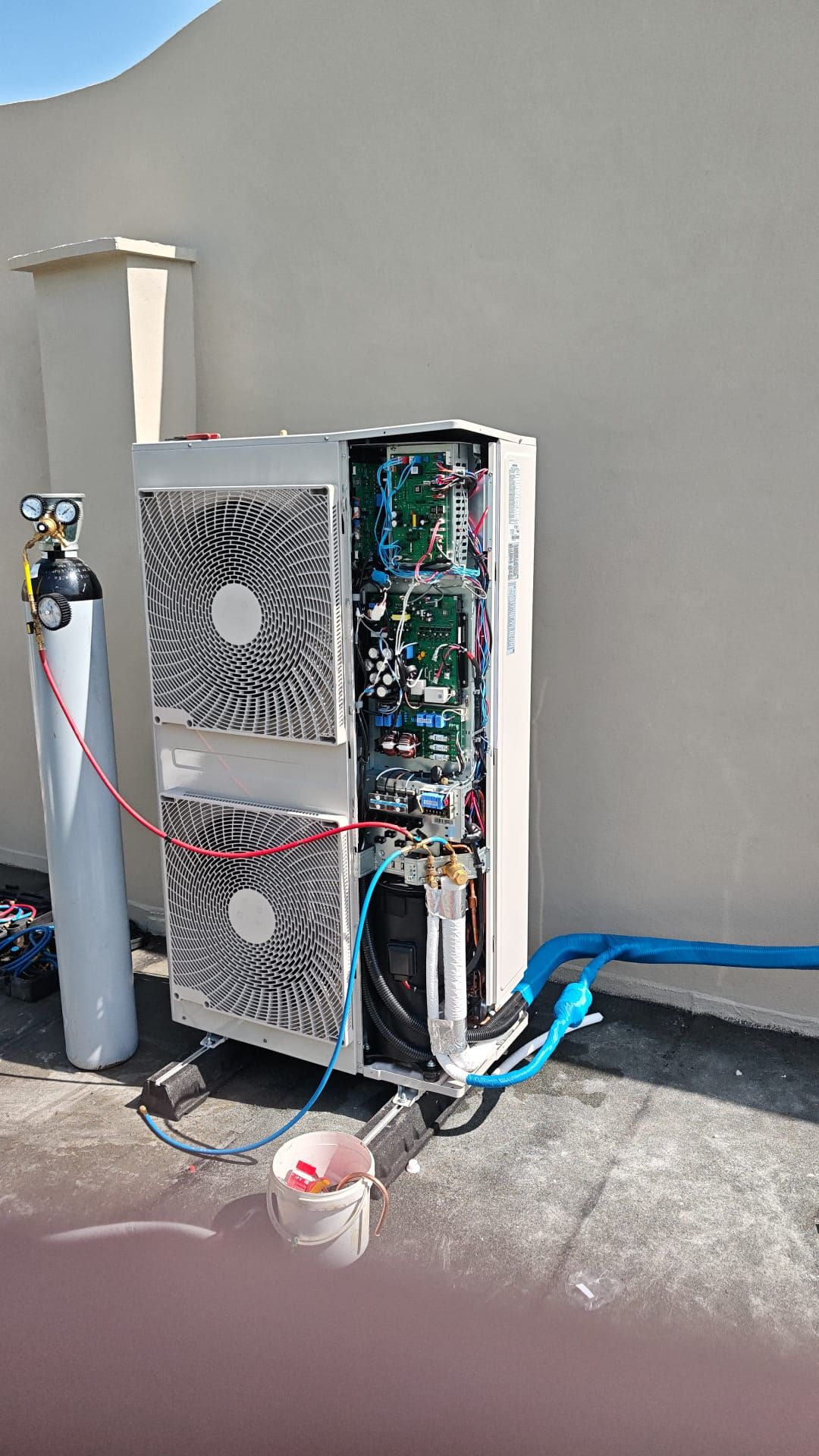 Unità HVAC esterna aperta, con cavi elettrici e tubi blu a vista, accanto a una parete e a una bombola del gas