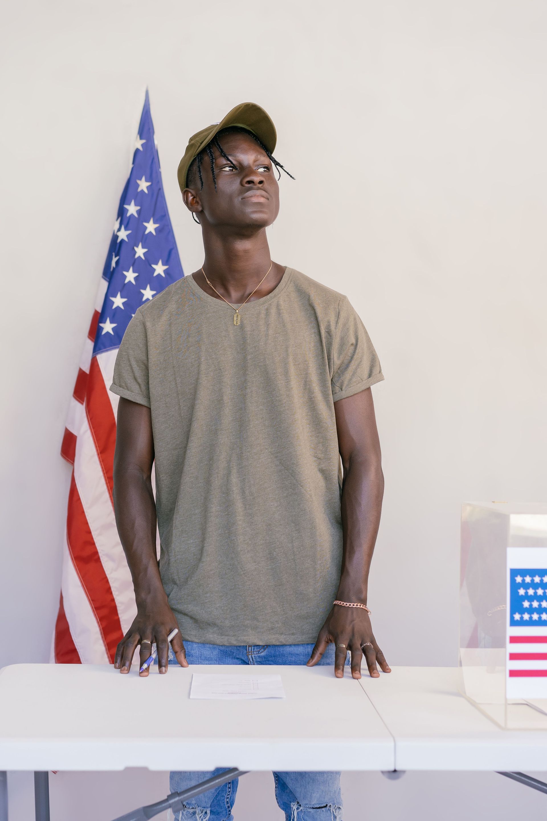 Man standing behind a table beside an American flag, looking upward in a plain room.